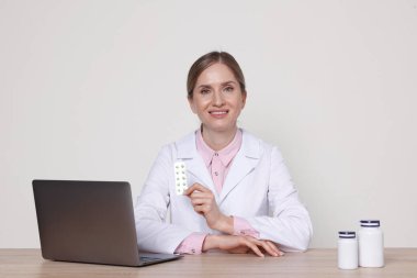 Palto giymiş gülümseyen eczacının portresi beyaz arka planda ilaç, dizüstü bilgisayar ve tıbbi şişeler ile ahşap masada