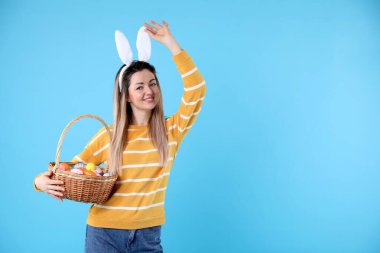 Paskalya kutlaması. Saç bandı takmış, tavşan kulaklı ve açık mavi arka planda boyalı yumurta dolu sepetli gülümseyen bir kadın.