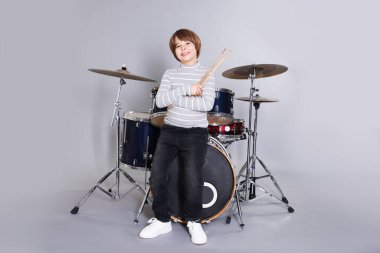 Açık gri arka planda bagetleri ve davulları olan küçük bir çocuk.