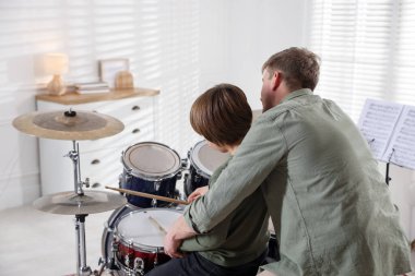 Küçük çocuk bateri çalmayı öğreniyor. İçeride müzik öğretmeniyle, arka planda.