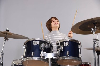 Açık gri arka planda bagetleri ve davulları olan küçük bir çocuk.