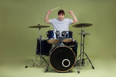 Beyzbol şapkalı gülümseyen genç, bagetleriyle yeşil arka planda davul çalmayı öğreniyor.