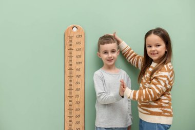 Küçük bir çocuk ve kız açık yeşil duvarın yanında boylarını grafikle ölçüyorlar.