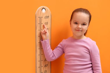 Küçük kız turuncu duvarın yanında boyunu ölçüyor.
