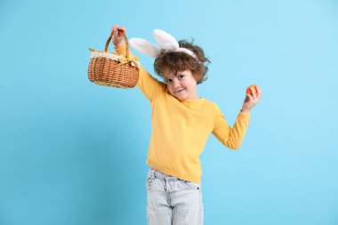 Paskalya kutlaması. Saç bantlı, tavşan kulaklı ve açık mavi arka planda boyalı yumurtalarla dolu sepeti olan sevimli bir çocuk.