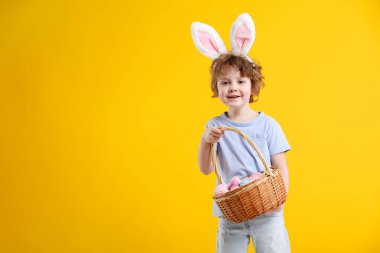 Paskalya kutlaması. Saçbantlı, tavşan kulaklı ve sarı arka planda boyalı yumurta dolu sepeti olan gülümseyen küçük bir çocuk. Metin için boşluk