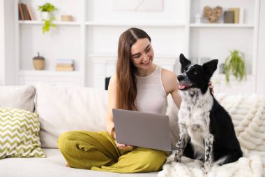 Evdeki kanepede dizüstü bilgisayarı ve sevimli köpeğiyle gülümseyen kadın.