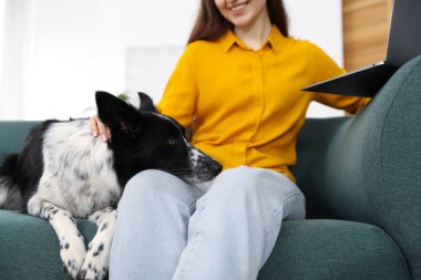 Evdeki kanepede dizüstü bilgisayarı ve sevimli köpeğiyle gülümseyen bir kadın.