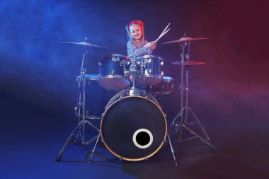 Bandanalı küçük bir kız karanlık arka planda davul çalmayı ve sigara içmeyi öğreniyor