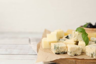 Serving board with different types of cheese and basil on light wooden table, closeup. Space for text