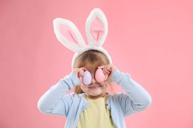 Paskalya kutlaması. Saç bandı takmış, tavşan kulaklı, kahverengi arka planda boyalı yumurtalarla gözlerini kapatan tatlı bir kız.