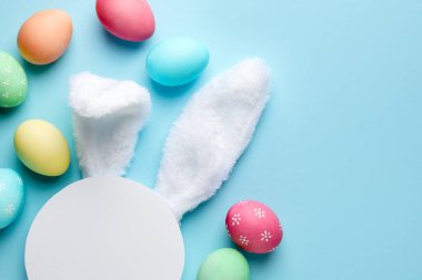 Mutlu Paskalyalar. Şirin tavşan kulakları, boş podyum ve açık mavi arka planda boyanmış yumurtalar, düz yatak. Metin için boşluk
