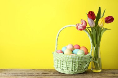 Hasır sepete boyanmış Paskalya yumurtaları ve ahşap masada sarı arka planda güzel laleler, metin için yer.