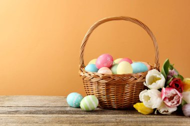 Hasır sepete boyanmış Paskalya yumurtaları ve ahşap masada soluk kahverengi arka planda güzel laleler.