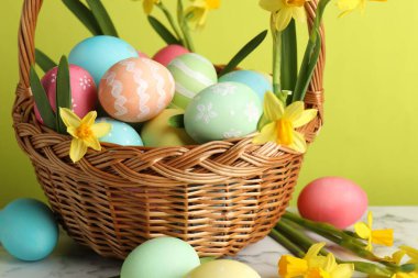 Boyanmış Paskalya yumurtaları ve hasır sepetin içindeki güzel nergisler açık yeşil arka planda beyaz mermer masa üzerinde, yakın plan