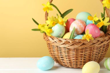 Boyanmış Paskalya yumurtaları ve hasır sepetin içindeki güzel nergisler bej arka planda beyaz ahşap masa üzerinde, yakın plan. Metin için boşluk