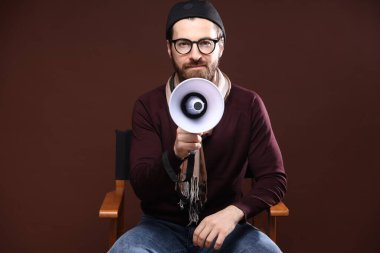 Kahverengi arka planda sandalyesinde megafonu olan profesyonel film yönetmeni.