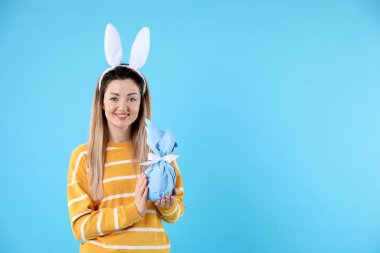 Paskalya kutlaması. Saçbandı takmış, tavşan kulaklı, açık mavi arka planda hediyesini tutan gülümseyen bir kadın. Mesaj için yer var.