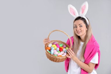 Paskalya kutlaması. Saç bandı takmış, tavşan kulaklı, gri arka planda yumurta dolu sepetli gülümseyen bir kadın. Mesaj için yer var.