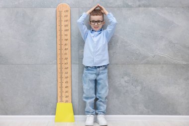 Boyunu grafikle ölçen küçük çocuk gri karo duvarın yanında.