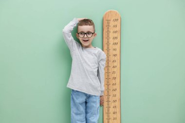 Küçük çocuk, açık yeşil duvarın yanında çizelgesiyle boyunu ölçüyor.