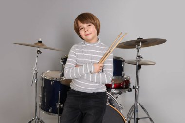 Açık gri arka planda bagetleri ve davulları olan küçük bir çocuk.