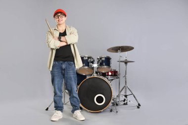 Gri arka planda davul seti ve bagetleriyle gözlüklü ve şapkalı gülümseyen genç çocuk.