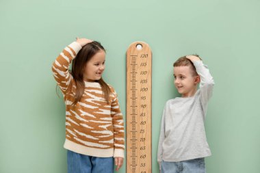 Küçük bir çocuk ve kız açık yeşil duvarın yanında boylarını grafikle ölçüyorlar.