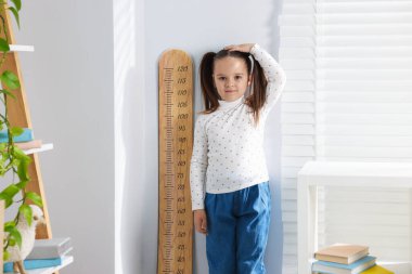 Küçük kız, evin içinde ışık duvarının yanında boyunu ölçüyor.