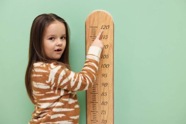 Açık yeşil duvarın yanında boyunu grafikle ölçen küçük bir kız.
