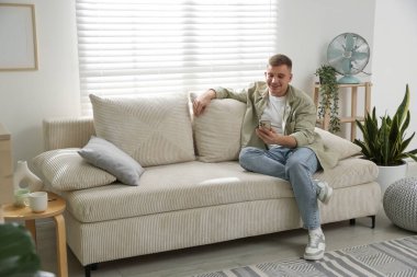 Akıllı telefonlu genç adam evdeki kanepede dinleniyor.