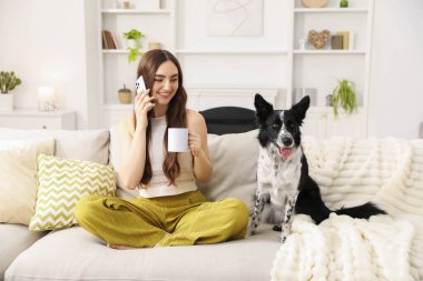 Elinde kupayla gülümseyen kadın akıllı telefonuyla ve evdeki kanepede sevimli köpeğiyle konuşuyor.