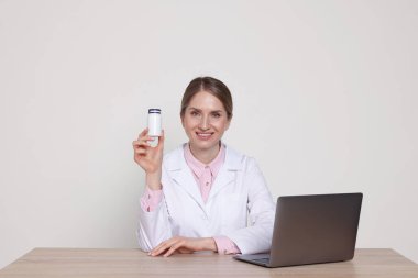 Beyaz arka planda, ahşap masada tıbbi şişe ve dizüstü bilgisayarla gülümseyen eczacının portresi.