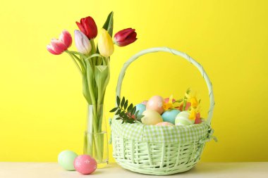 Hasır sepete boyanmış Paskalya yumurtaları ve sarı arka planda bej masadaki güzel laleler.