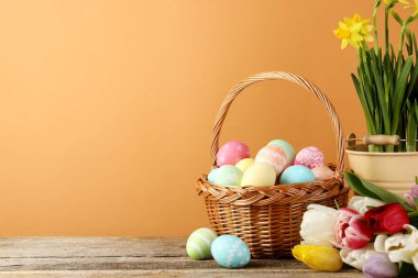 Hasır sepete boyanmış Paskalya yumurtaları ve ahşap masada soluk kahverengi arka planda güzel çiçekler, yakın plan. Metin için boşluk