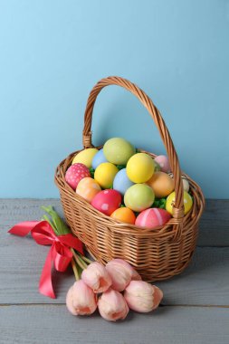 Hasır sepete boyanmış Paskalya yumurtaları ve açık mavi arka plana karşı gri ahşap masada güzel laleler.