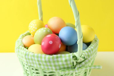 Hasır sepetin içine boyanmış Paskalya yumurtaları beyaz masada sarı arka planda, yakın plan.