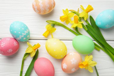 Painted Easter eggs and beautiful daffodils on white wooden table, flat lay