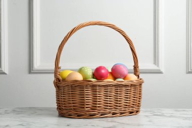 Hasır sepete boyanmış Paskalya yumurtaları beyaz duvarın yanındaki beyaz mermer masada.