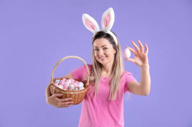 Paskalya kutlaması. Saç bantlı, tavşan kulaklı ve menekşe arkaplanda boyalı yumurtalarla dolu sepeti olan gülümseyen bir kadın.