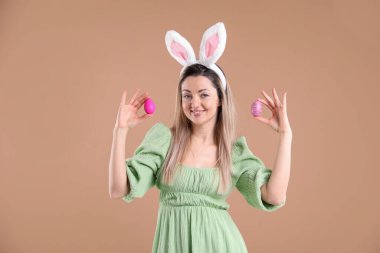 Paskalya kutlaması. Bej arka planda boyalı yumurta tutan, saç bandı takmış, tavşan kulaklı, gülümseyen bir kadın portresi.