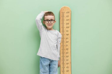 Küçük çocuk, açık yeşil duvarın yanında çizelgesiyle boyunu ölçüyor.