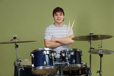 Beyzbol şapkalı, davul setiyle yeşil arka planda gülümseyen genç çocuk.