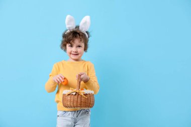 Easter celebration. Smiling little boy in headband with bunny ears and wicker basket full of painted eggs on light blue background. Space for text
