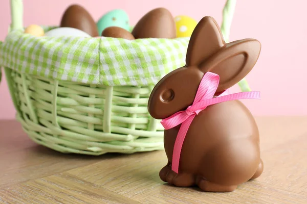 Çikolatalı Paskalya tavşanı ve hasır sepetteki yumurtalar ahşap masada pembe arka planda, yakın plan.