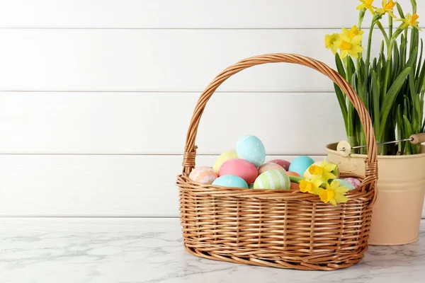 Hasır sepete boyanmış Paskalya yumurtaları ve beyaz mermer masaya güzel nergisler, yazı için yer.