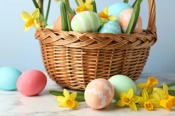 Boyanmış Paskalya yumurtaları ve hasır sepetin içindeki güzel nergisler beyaz mermer masa üzerinde açık mavi arka plan, yakın plan