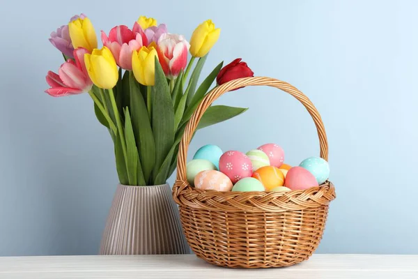 Hasır sepetin içine boyanmış Paskalya yumurtaları beyaz ahşap masa üzerinde ve açık mavi arka planda vazoda güzel laleler.