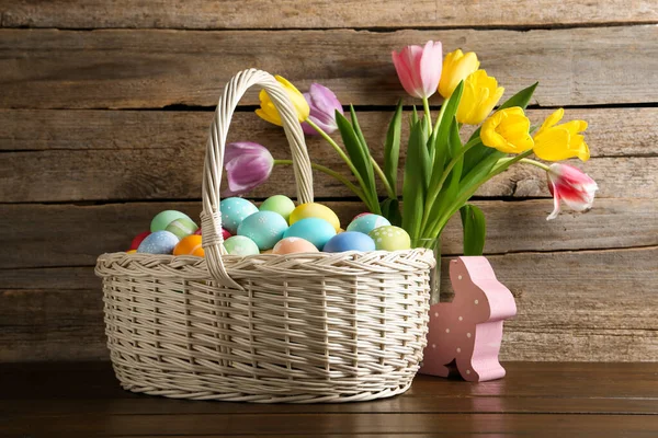 Hasır sepete boyanmış Paskalya yumurtaları, dekoratif tavşan figürü ve ahşap masada güzel laleler.