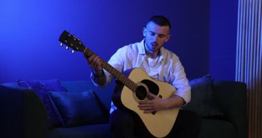 Gitar çalan ve mavi ışıkta şarkı söyleyen bir adam. Kamera sağa hareket ediyor